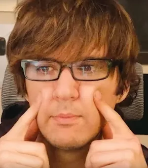 Ruben Sim wearing prescription glasses with his index fingers touching either side of his cheeks.